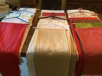 Photo showing multiple rectangular tablecloths in colors including red, beige, cream, and white, folded and laid out with hangers on top