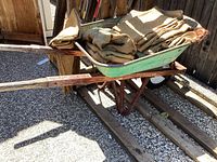 Side and angled view of vintage metal wheelbarrow containing folded burlap sacks, showing rusty areas and wooden handles.