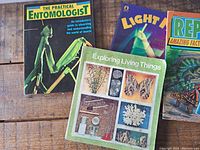 Top view of 4 science books, including The Practical Entomologist, Light Magic, Exploring Living Things, and a Reptiles facts book.