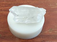 Clear quartz crystal resting on a white round stand on a wooden surface. Crystal appears translucent with natural formations.