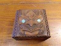 View of the ornate carved lid of the wooden trinket box showing the detailed face motif with inlaid clear eyes and decorative border carvings.