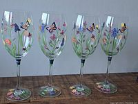 Four clear glass goblets with colorful hand painted floral and butterfly designs, arranged side-by-side on a wooden surface.