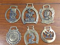 Overview of six brass horse brasses arranged on wood surface. Shows shape, size, and detail variety.