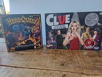 Photo of two board game boxes side by side on a wooden surface. Left is Hero Quest Mage of the Mirror quest pack, right is Clue Liar's Edition.