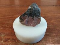 Raw Rhodonite stone displayed on a white cylindrical stand on a wooden surface, showing its pink and black coloration and rough texture.