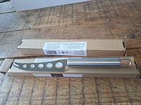 Two cheese cutting knives laid on a wooden surface with their cardboard boxes underneath, showing the knives' full blade and handle design.