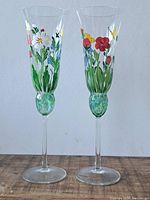 Pair of two clear flute glasses with colorful hand-painted floral designs on the bowls and decorative greenish stems.
