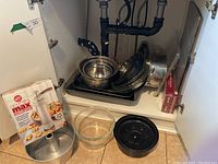 Photo showing assorted baking and roasting pans arranged under a sink including nested stainless steel bowls, baking and roasting pans, fluted cake pan, glass bowl, and Betty Crocker Cookie Max cookie press kit in box.