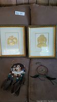 Photo of the two framed gold leaf impressions by Bill Reid alongside a dream catcher and a miniature drum decoration, all displayed on a couch.