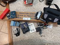 Full overview of the lot showing Ricoh AF-50 camera with case and manual, JVC Everio camcorder with case, projection screen box, assorted camera accessories including flashes and cables.