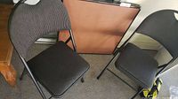 Two black cushioned folding chairs with metal frames on carpeted floor.
