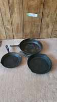 Three cast iron frying pans arranged on carpeted floor, showing the largest Wagner brand pan and two smaller pans, all with used surfaces.