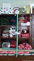 Full view of wooden cabinet shelves holding assorted Christmas-themed towels, oven mitts, gift boxes, plush toy, and wrapping paper on lower shelf.