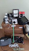 Photo showing multiple electronic devices on two wooden nightstands including Panasonic cordless phones, Sylvania digital photo frame, LG DVD player, small speakers, webcam in packaging, and Sunbeam portable heater on floor with cords
