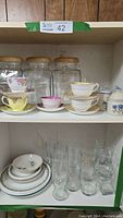 Shelf showing five china cups and saucers with floral and gold trim design on top shelf. Below, some Corningware plates and various clear glass beer and wine glasses.