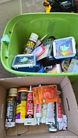 Green plastic bin and cardboard box containing tubes of adhesives, sealants, and small boxed hardware items.