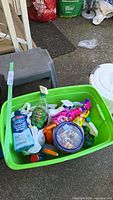 Photo showing green plastic bin with assorted cleaning supplies, gray two-step stool, and small white rubbish bin with lid.