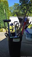 Bucket filled with various longhandled yard tools including rakes, shovels, broom, pitchfork, and sprinkler nozzle.