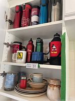Overview of upper kitchen cabinet shelf filled with multiple stainless steel to-go mugs, pottery mugs, vintage plates and bowls, a glass stein, and a red insulated jug cover with Mickey Mouse graphic.