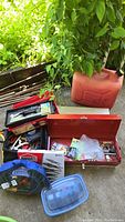 Photo showing two steel tool boxes, a black tool bag filled with hand tools, a red fuel container, and several plastic containers with hardware and tools on concrete ground under outdoor greenery.