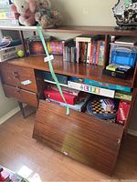 Front view of vintage wooden sideboard showing three drawers, drop-down door open revealing interior storage space with board games and other miscellaneous items inside, top shelf with books and figurines.