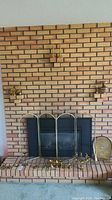 Wide view of fireplace with brick surround and several brass and copper fireplace accessories including multi-panel brass fire screen, candle holders, fire tools, and bellows.