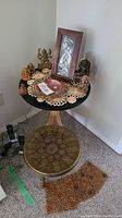 Full view of brass stool in front of a small round wooden table with various figurines, framed artwork, and brass bowl on top.