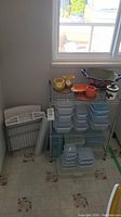 Photo shows metal wire rack with multiple plastic Food Saver containers stacked on shelves, along with assorted dishes on top. Rack missing a corner cap piece.
