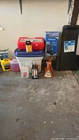 View showing hydraulic jack, jack stands, automotive creeper, red plastic case with bottle jacks, orthopedic cushion box, and assorted automotive liquids.