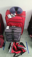 Lot includes a large red soft-sided rolling suitcase with a travel neck pillow on top, and a smaller grey soft-sided rolling suitcase with strap in front.