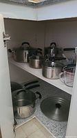 Photo showing lower and upper shelves of a cabinet with multiple stainless steel pots and pans with black handles, accompanied by a clear glass measuring cup with markings.