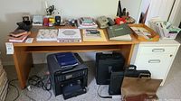Photo showing large wooden desk with printer, notebooks, binders, pens, stapler, and briefcases.