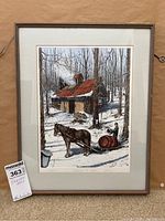 Framed signed winter cabin art print hanging vertically on a wall.