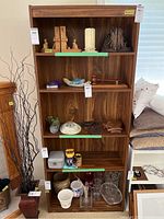 Front view of tall manufactured wood bookshelf filled with various decorative items such as wooden figurines, a ceramic dish, glassware and small plants. Shelves are solid and finish is dark wood grain.