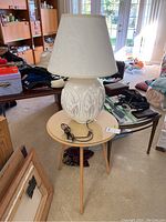 White ceramic lamp with floral relief design, large white floral lampshade, and brown power cord; placed on round accent table with wood legs and glass top in interior room setting.
