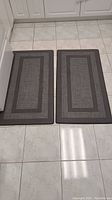 Pair of two gray chef's mats placed side by side on white tiled floor, showing full mats and floor context.