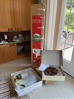 Six-foot artificial Christmas tree box upright next to two wreath boxes on carpeted floor near glass door and wooden cabinet.