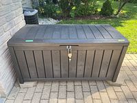 Front view of brown resin deck box showing woodgrain panel style and padlock with keys attached