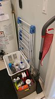 Folded white and blue clothes drying rack, plastic laundry basket with cleaning bottles, and brown storage bin with additional supplies in corner near water heater.