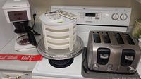 Kitchen appliances and serveware arranged on kitchen stove and counter: Bunn coffee maker, stackable white pie rack, glass cake stand, Hamilton Beach 4-slice toaster.