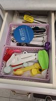Top view inside drawer showing various measuring spoons, peelers, spatulas, and other small kitchen gadgets in plastic baskets.