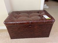 Front view of brown faux leather padded storage bench with button tufting on top and label on lid.