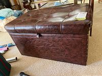 Top and side view of the brown faux leather padded storage bench seat showing tufted cushion top and nailhead trim, placed on carpet with household items in background.