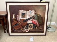 Framed oil painting on canvas showing a still life of violin, books, candle, and other objects on a table.