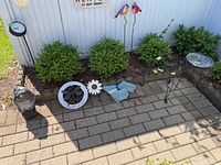 Wide view of outdoor garden items including metal plant stand, resin stones, metal flower, plastic owl, metal birds, and garden clock placed on ground outdoors.