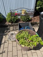 View of the full lot showing metal plant stand, resin planter with plants, blue hose, plastic lid, and red decorative items
