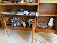 Full view of wooden cabinet with three shelves containing assorted servingware items such as trays, platters, and a white water jug.