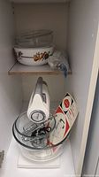 View inside cabinet shelf showing additional glass bowls with lids and fruit patterned bowl, plastic-wrapped mixer accessories.