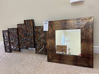 Photo showing the steel wall art with several vertical panels installed next to a square wall mirror with a textured metallic frame laid on the floor.