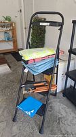 Black folding step ladder with BBQ utensils, blue tarp, and folded table covers arranged on steps.